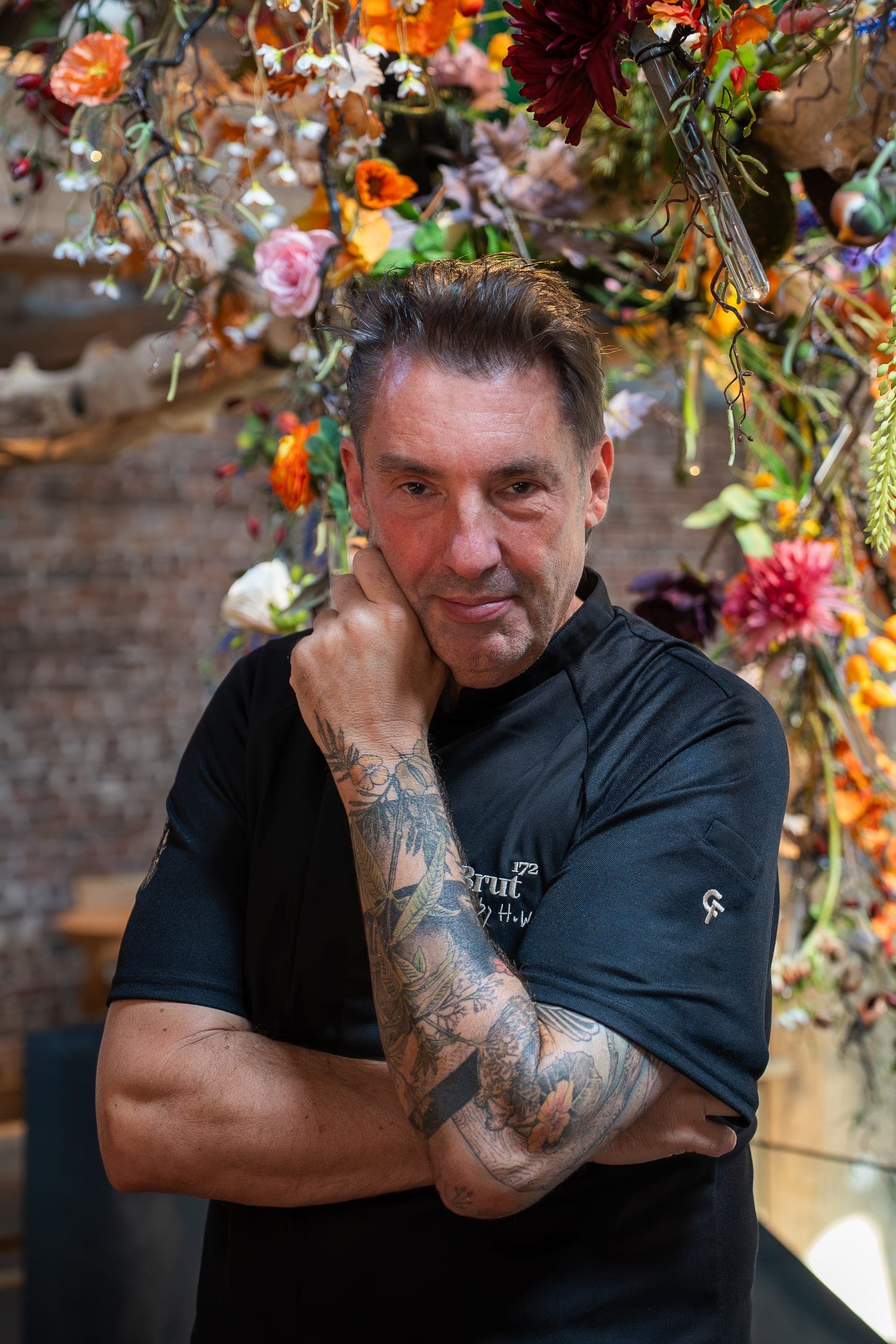Hans van Wolde in einer schwarzen Cheffix-Kochjacke, die Hand unter dem Kinn, vor einer Decke voller Blumen